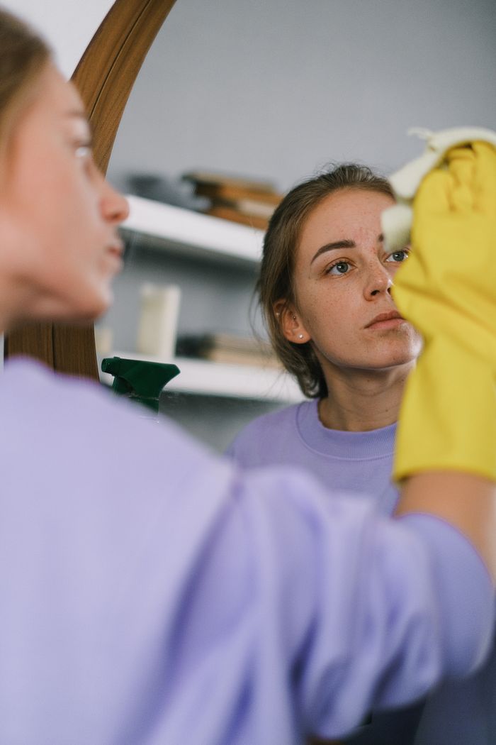 hero-img Crop female in purple sweatshirt and latex gloves washing mirror in light apartment in daytime