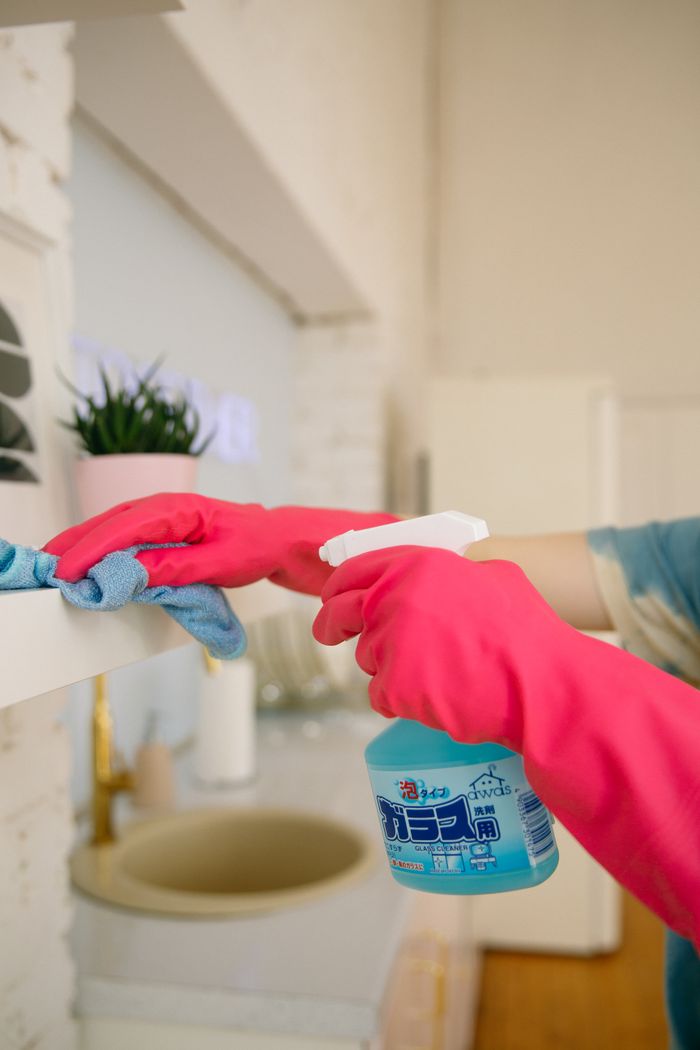 about-01 Person using spray bottle and cloth for cleaning a kitchen shelf, ensuring hygiene and cleanliness.