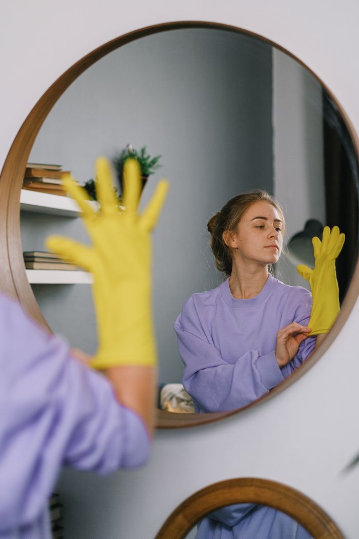 services-01 A woman wearing yellow gloves is reflected in a wall mirror indoors, exuding a serene expression.